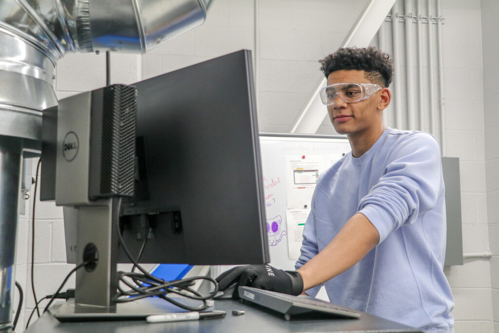 College student standing at a computer, looking at the screen and wearing safety goggles.