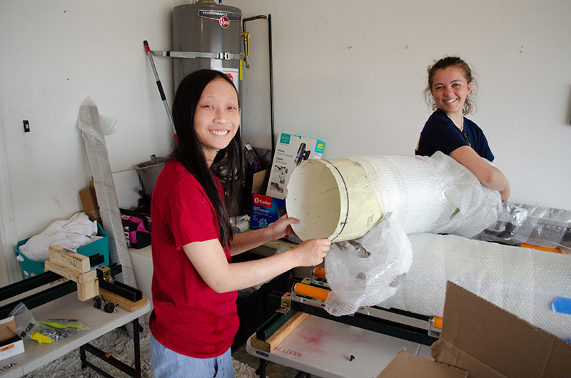 Two girls smiling at the camera while taking bubble wrap off of a package.