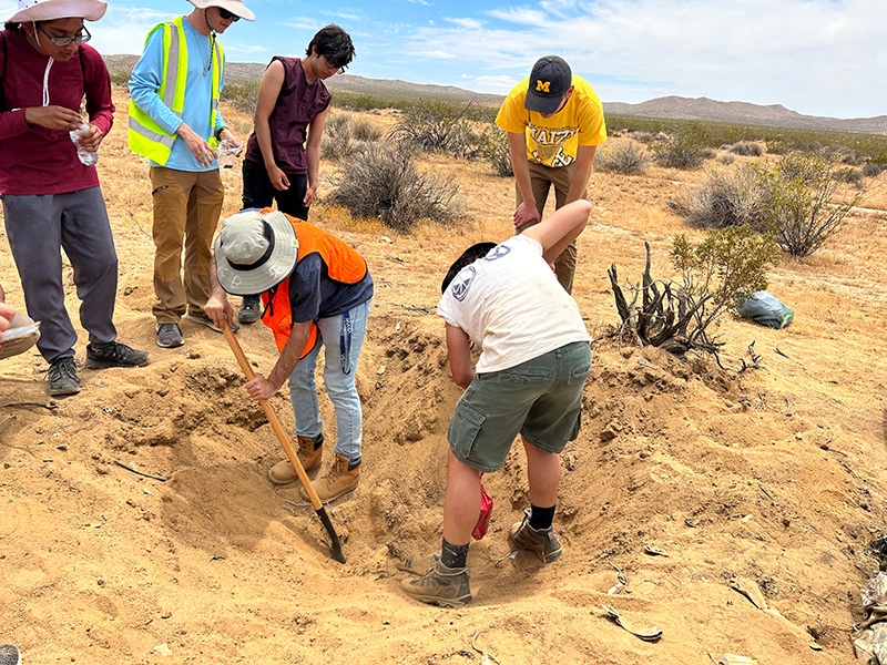 Two people digging in the desert to recover rocket debris with four other people standing around them.